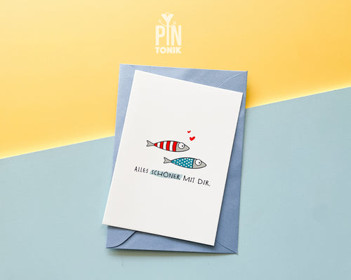 Premium postcard with an original illustration of two small fish (one red striped, one blue dotted) with tiny red hearts and the German text 'ALLES SCHÖNER MIT DIR.', shown with a luxury blue envelope.
