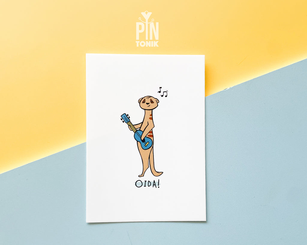Premium postcard with an original illustration of a meerkat playing a blue ukulele with the text 'OIDA!', shown against a split yellow and light blue background.