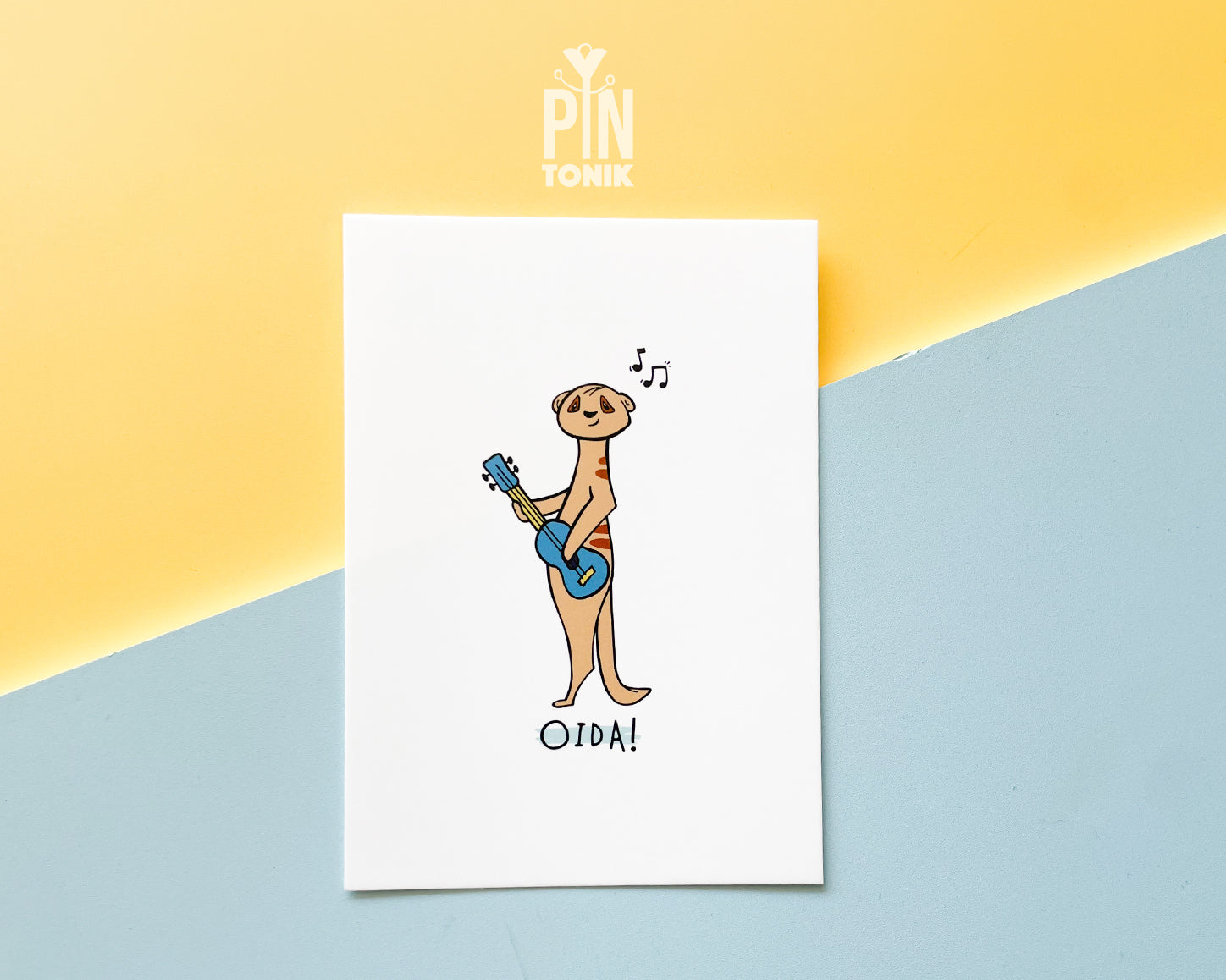Premium postcard with an original illustration of a meerkat playing a blue ukulele with the text 'OIDA!', shown against a split yellow and light blue background.