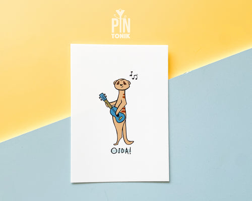 Premium postcard with an original illustration of a meerkat playing a blue ukulele with the text 'OIDA!', shown against a split yellow and light blue background.