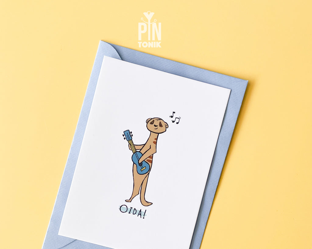 Premium postcard with an original illustration of a meerkat playing a blue ukulele with the text 'OIDA!', shown against a light blue background.