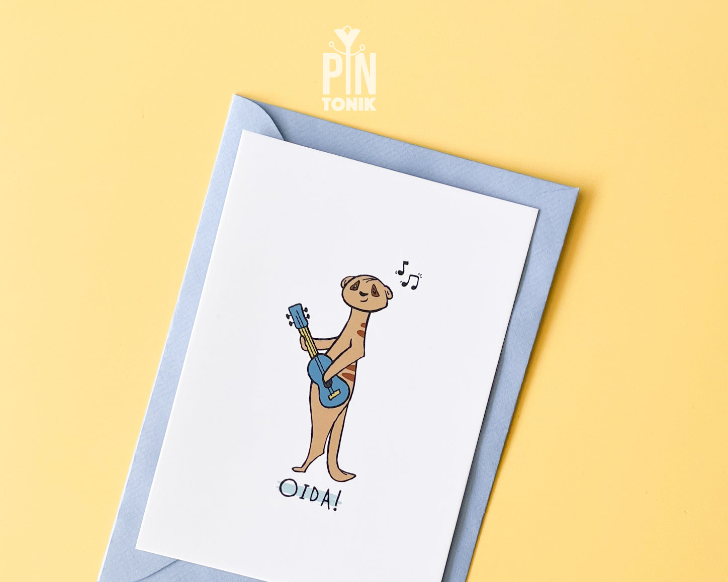 Premium postcard with an original illustration of a meerkat playing a blue ukulele with the text 'OIDA!', shown against a light blue background.