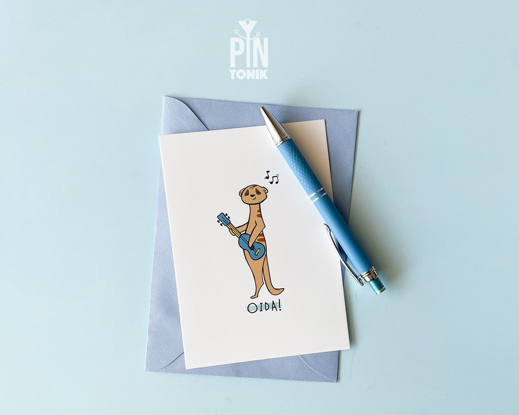 Premium postcard with an original illustration of a meerkat playing a blue ukulele with the text 'OIDA!', shown against a light blue background.