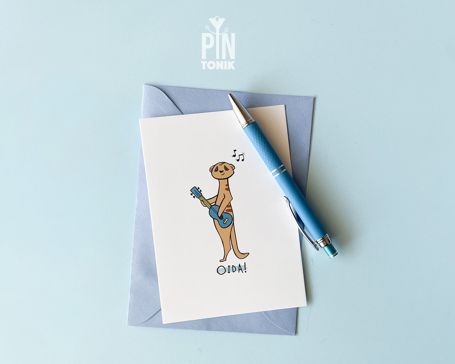 Premium postcard with an original illustration of a meerkat playing a blue ukulele with the text 'OIDA!', shown against a light blue background.