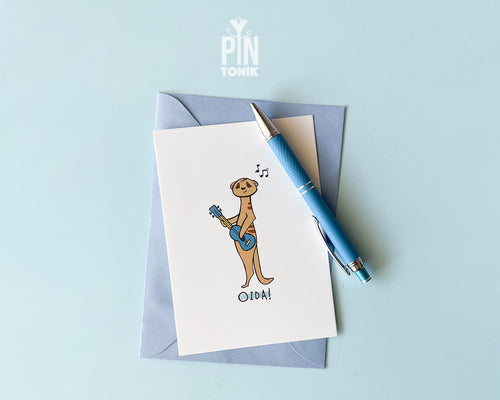 Premium postcard with an original illustration of a meerkat playing a blue ukulele with the text 'OIDA!', shown against a light blue background.