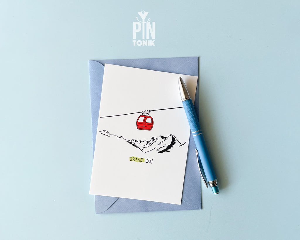 Premium postcard with an original illustration of a red cable car and the text 'Griaß Di!', shown against a blue background with a light blue envelope.