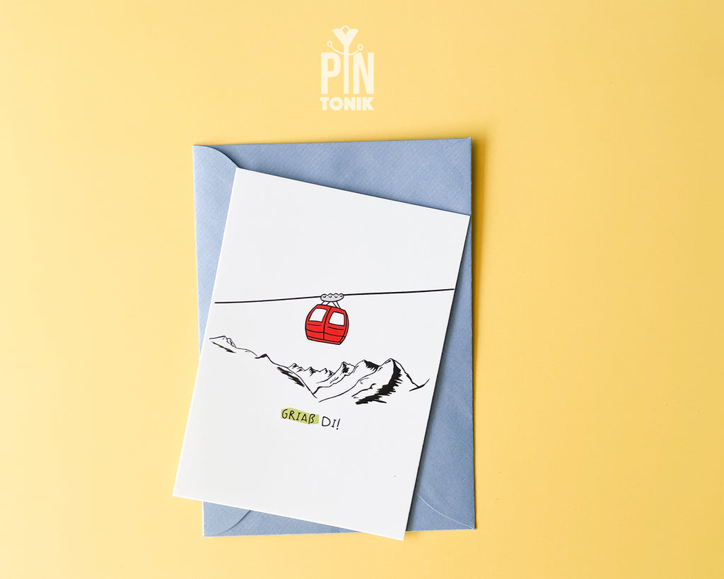 Premium postcard with an original illustration of a red cable car and the text 'Griaß Di!', shown against a vibrant yellow background with a light blue envelope.