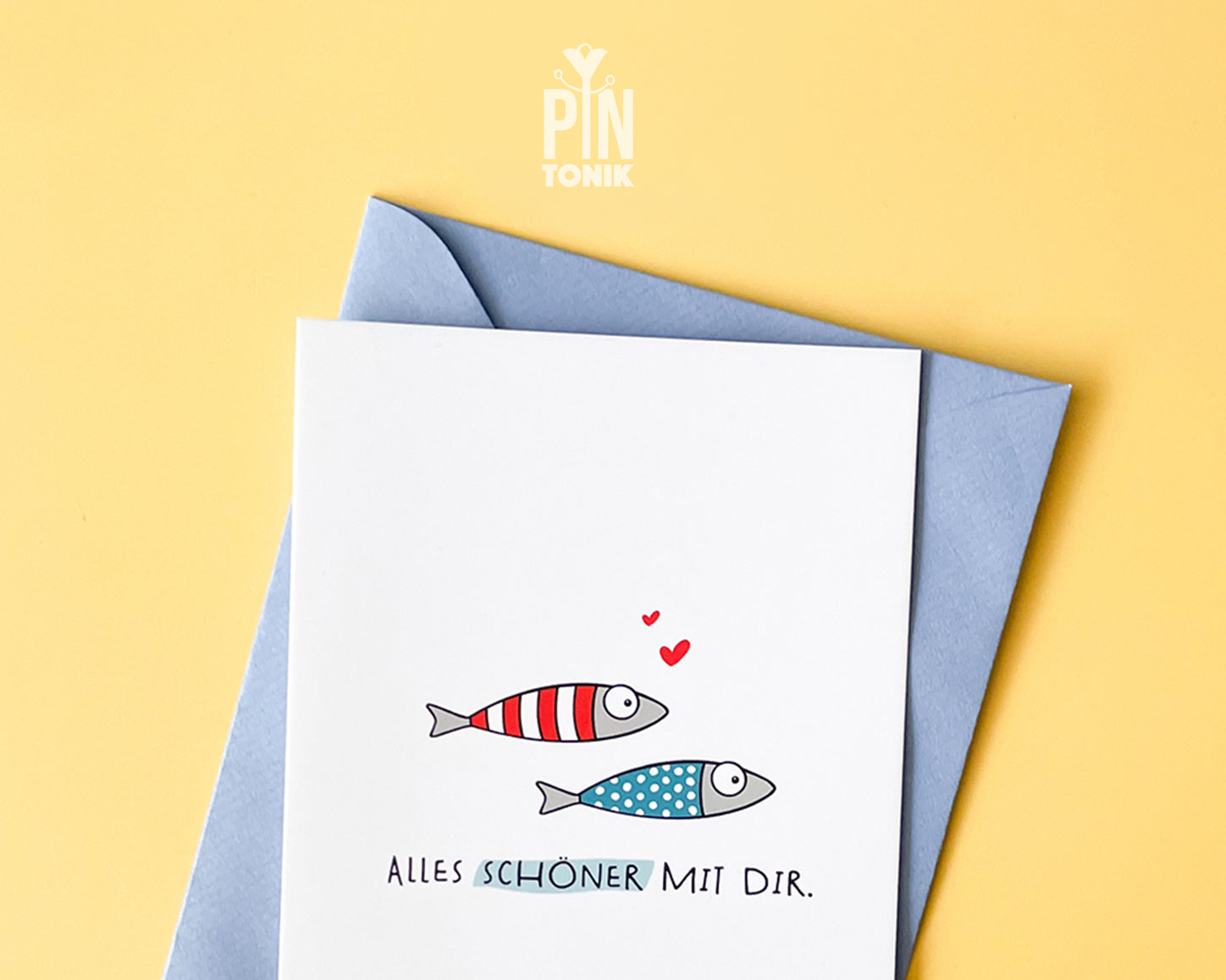 Premium postcard with an original illustration of two small fish (one red striped, one blue dotted) with tiny red hearts and the German text 'ALLES SCHÖNER MIT DIR.', shown with a luxury blue envelope.
