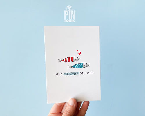 Premium postcard with an original illustration of two small fish (one red striped, one blue dotted) with tiny red hearts and the German text 'ALLES SCHÖNER MIT DIR.', shown with a luxury blue envelope.