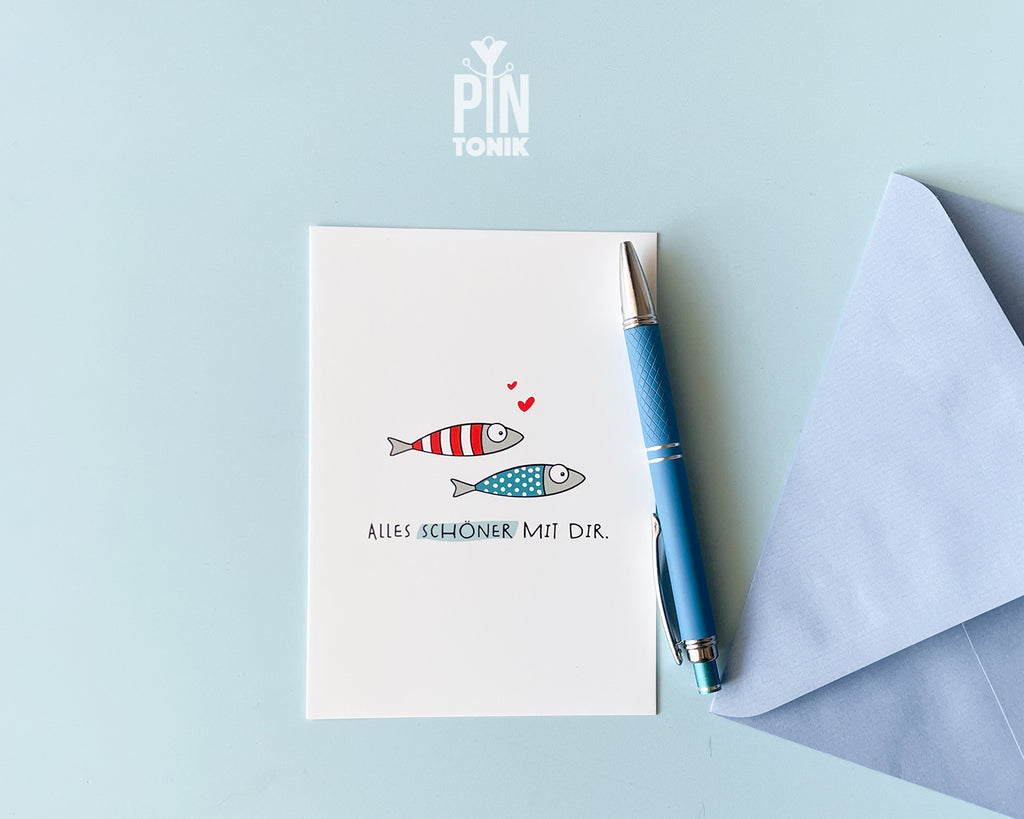 Premium postcard with an original illustration of two small fish (one red striped, one blue dotted) with tiny red hearts and the German text 'ALLES SCHÖNER MIT DIR.', shown with a luxury blue envelope.