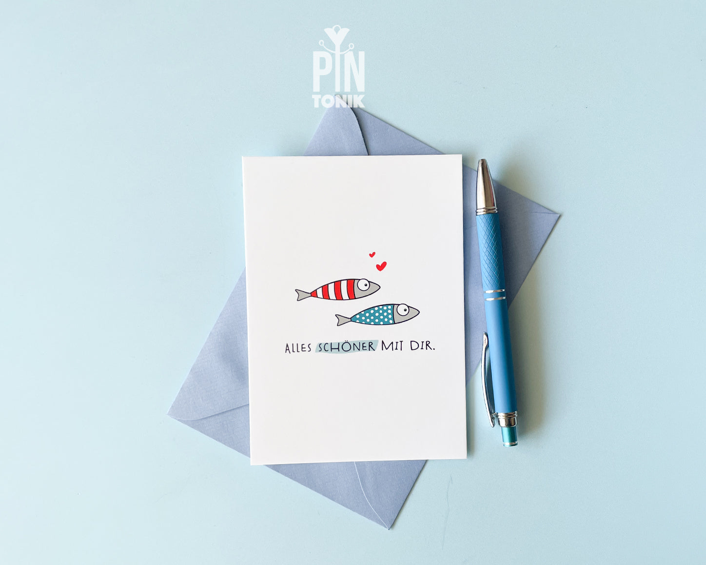 Premium postcard with an original illustration of two small fish (one red striped, one blue dotted) with tiny red hearts and the German text 'ALLES SCHÖNER MIT DIR.', shown with a luxury blue envelope.