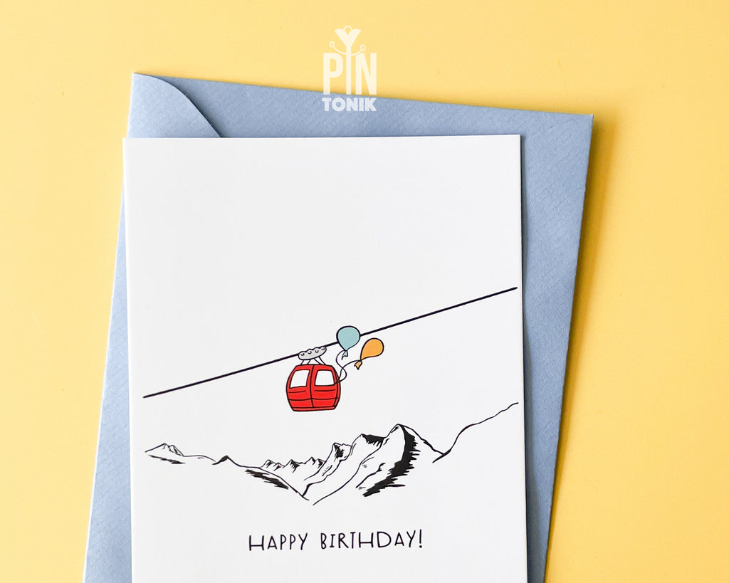 Premium postcard with an original illustration of a red cable car with colorful balloons and the text 'HAPPY BIRTHDAY!', shown against a yellow background with a light blue envelope.