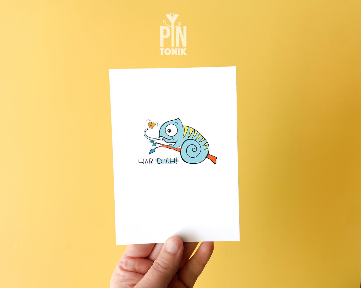 Premium postcard featuring Moji the Chameleon, the PinTonik mascot, catching a butterfly with the text 'HAB DICH!', shown with a luxury light blue envelope.