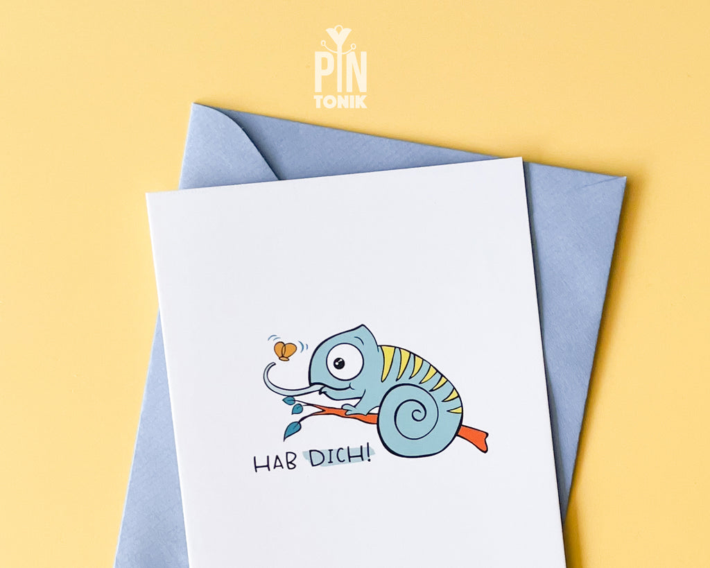 Premium postcard featuring Moji the Chameleon, the PinTonik mascot, catching a butterfly with the text 'HAB DICH!', shown with a luxury light blue envelope.