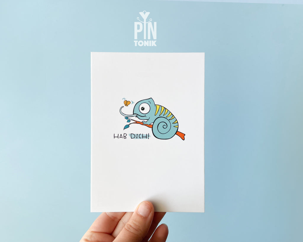 Premium postcard featuring Moji the Chameleon, the PinTonik mascot, catching a butterfly with the text 'HAB DICH!', shown with a luxury light blue envelope.