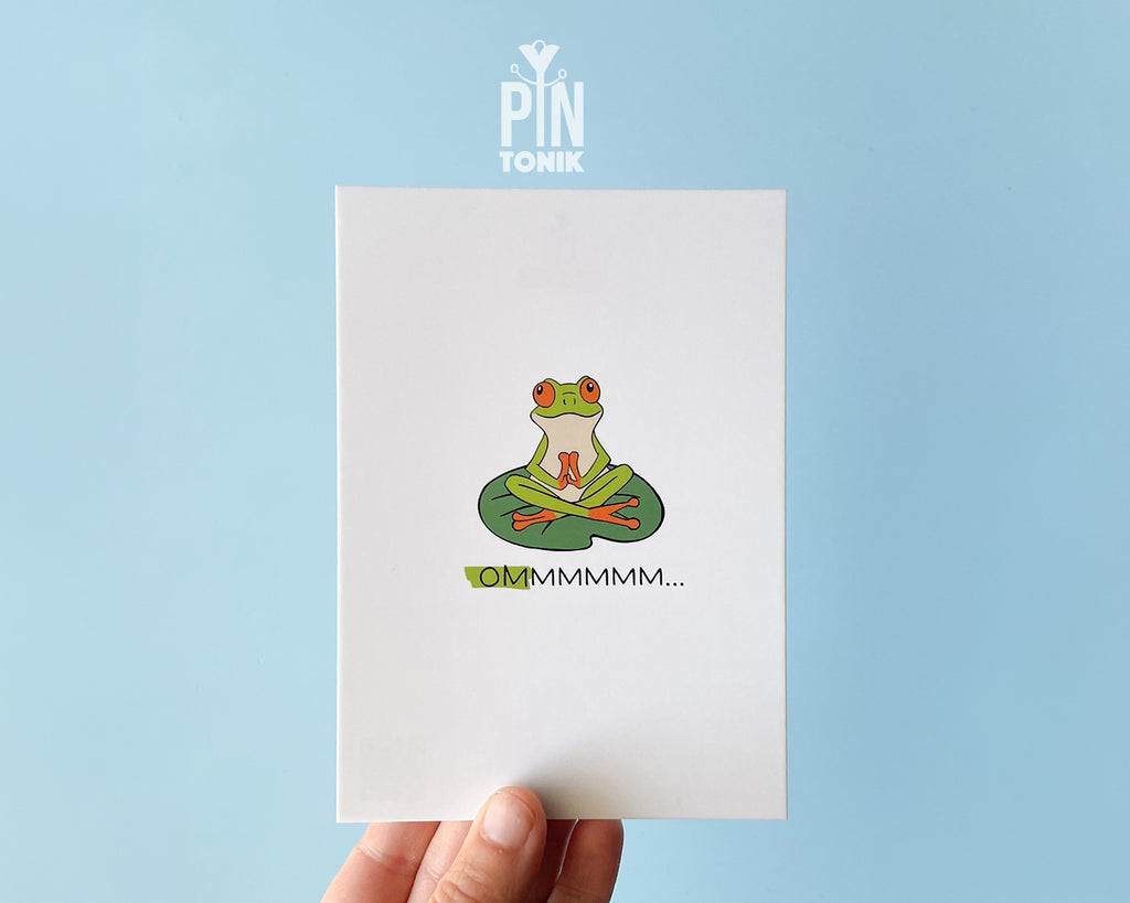 Premium postcard with an original illustration of a green tree frog meditating on a lily pad with the text 'OMMMMMM...', shown held against a light blue background.