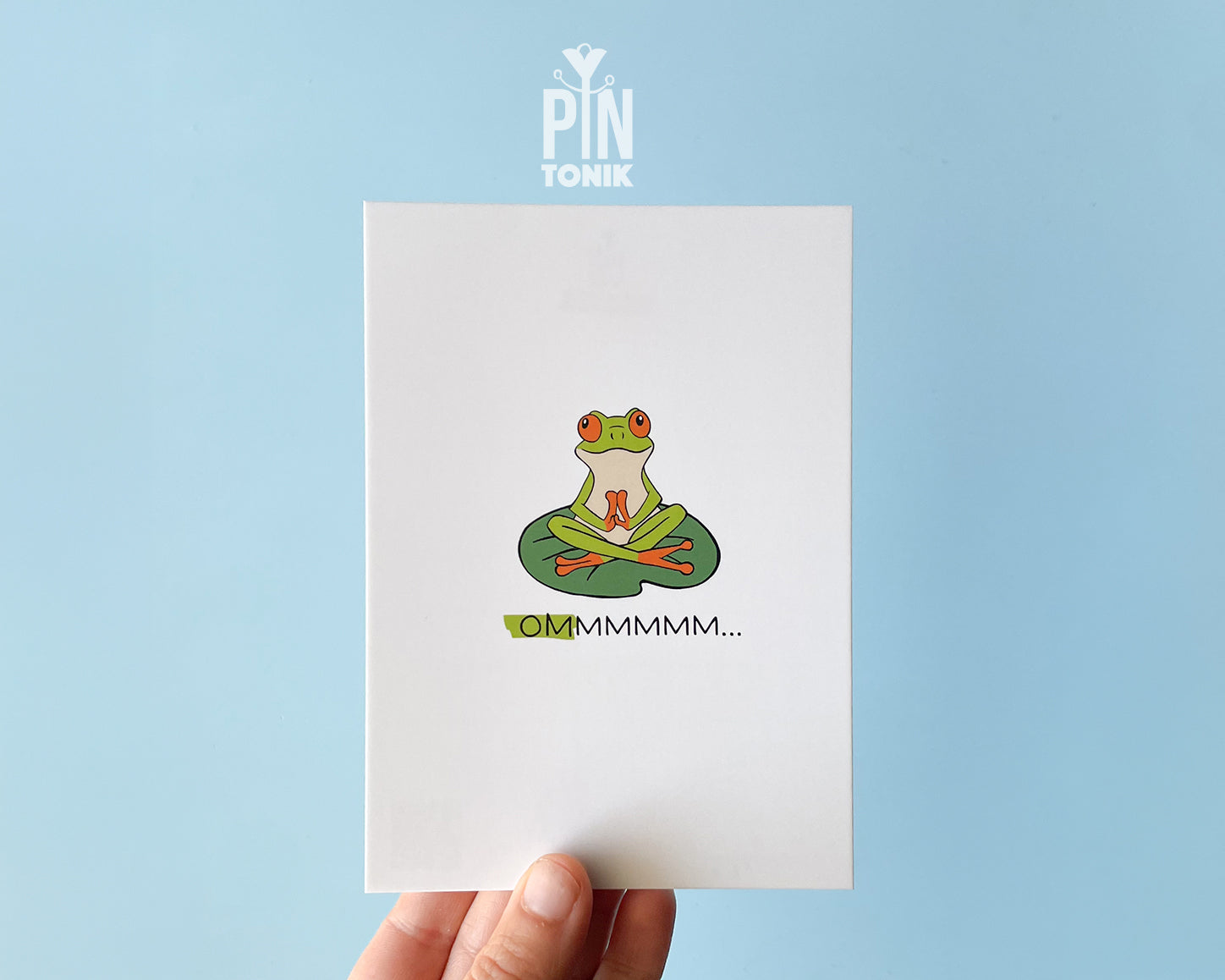 Premium postcard with an original illustration of a green tree frog meditating on a lily pad with the text 'OMMMMMM...', shown held against a light blue background.