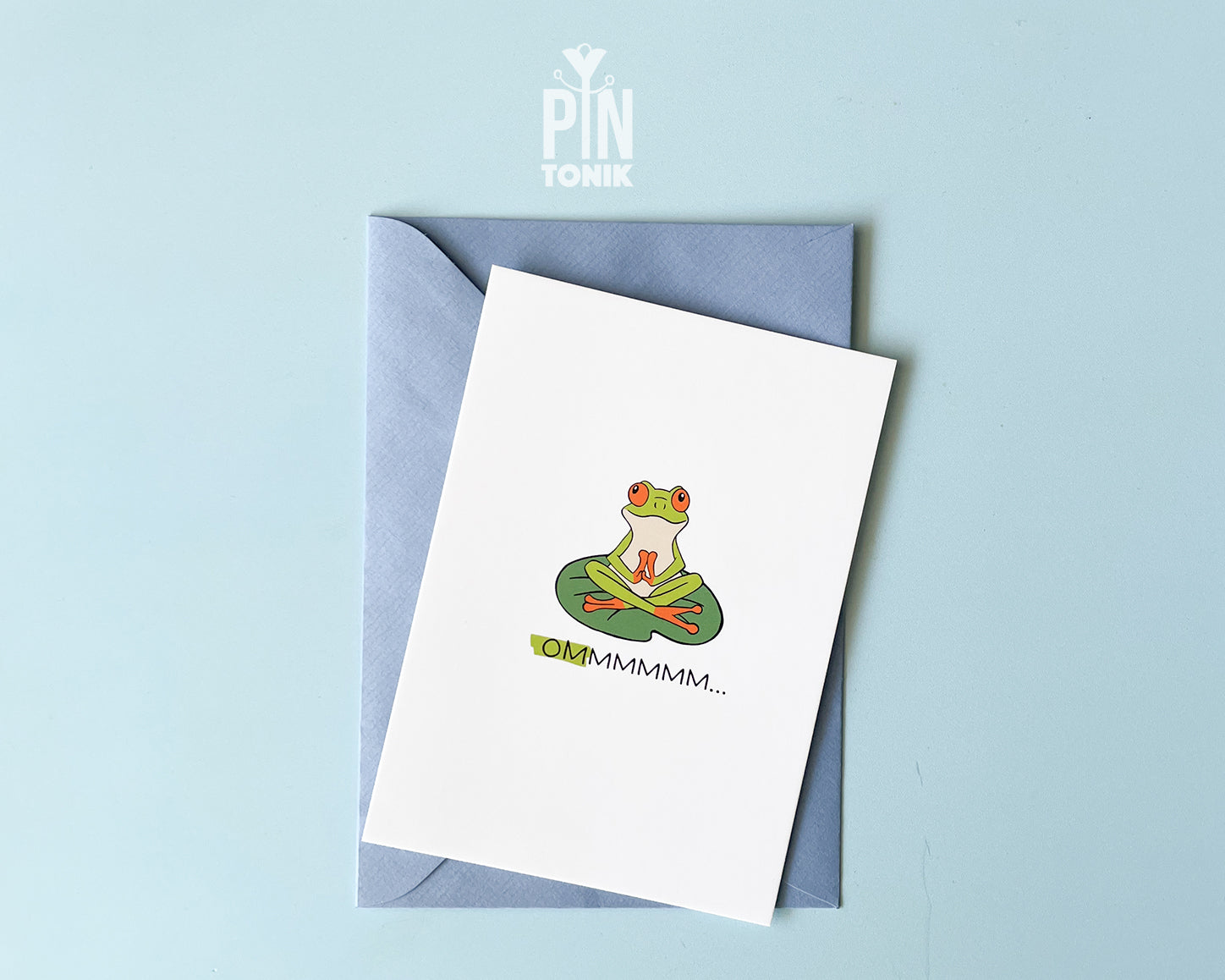Premium postcard with an original illustration of a green tree frog meditating on a lily pad with the text 'OMMMMMM...', shown held against a light blue background.