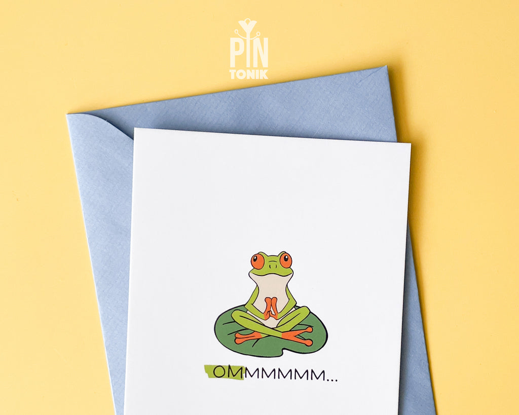 Premium postcard with an original illustration of a green tree frog meditating on a lily pad with the text 'OMMMMMM...', shown held against a light blue background.