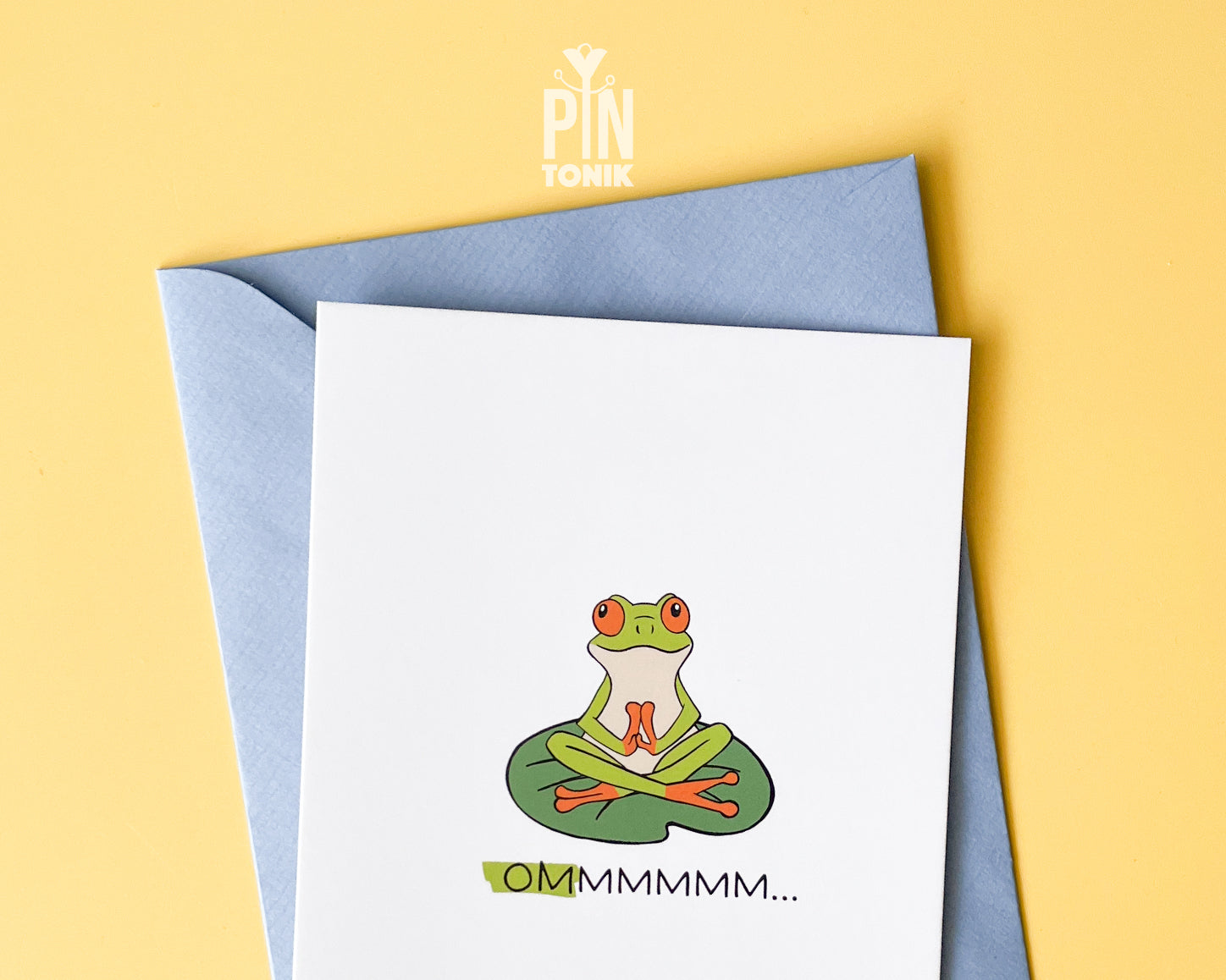 Premium postcard with an original illustration of a green tree frog meditating on a lily pad with the text 'OMMMMMM...', shown held against a light blue background.