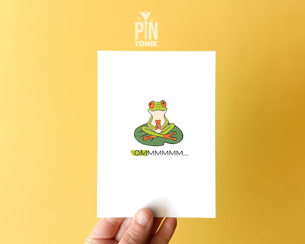 Premium postcard with an original illustration of a green tree frog meditating on a lily pad with the text 'OMMMMMM...', shown held against a light blue background.