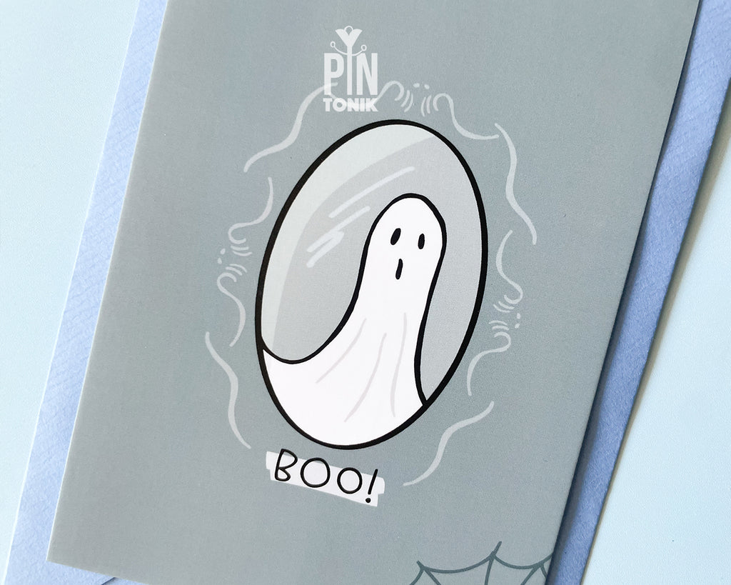 Premium grey postcard with an original illustration of a ghost in a mirror and the text 'BOO!', shown with a light blue luxury envelope on a light blue background.