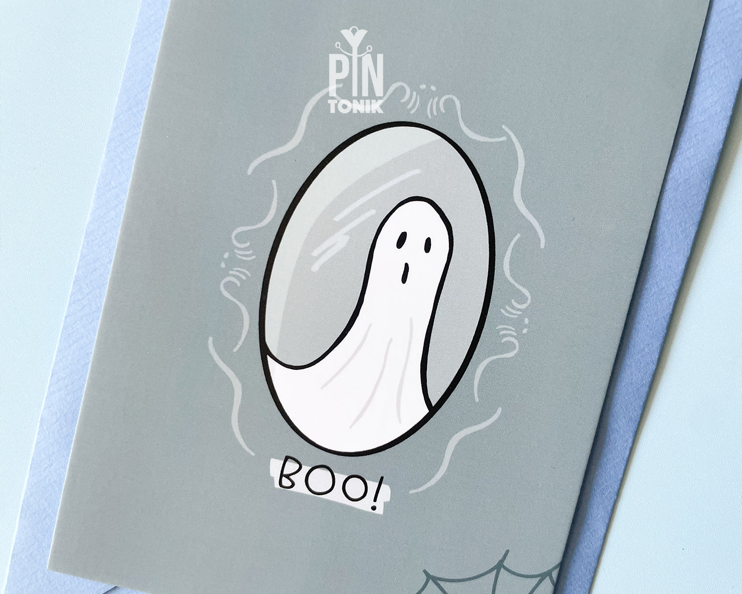 Premium grey postcard with an original illustration of a ghost in a mirror and the text 'BOO!', shown with a light blue luxury envelope on a light blue background.