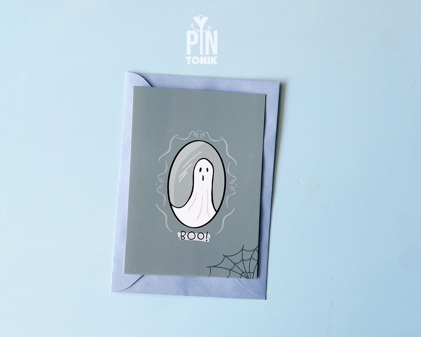 Premium grey postcard with an original illustration of a ghost in a mirror and the text 'BOO!', shown with a light blue luxury envelope on a light blue background.