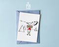Premium postcard with an original illustration of a mountain gnome holding a 'SERVUS' sign in a snowy alpine landscape, shown with a light blue envelope.