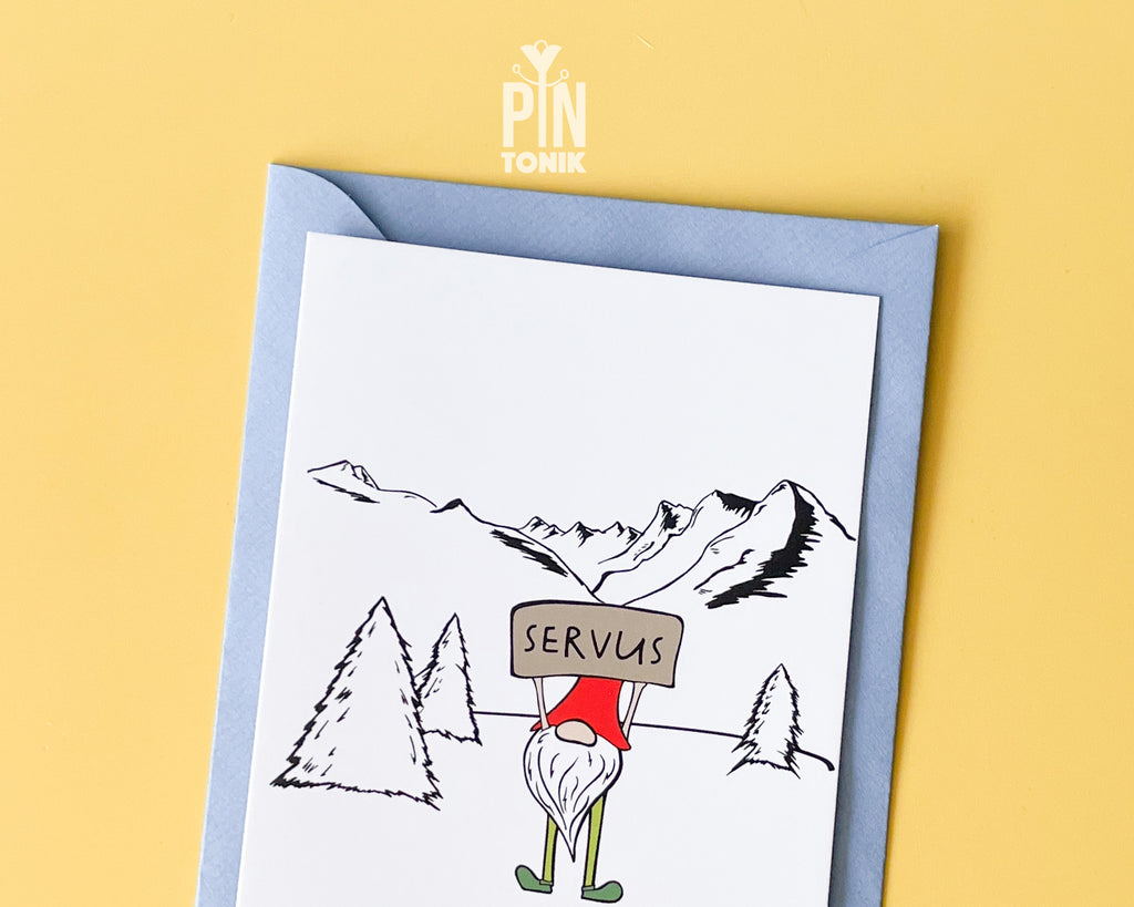 Premium postcard with an original illustration of a mountain gnome holding a 'SERVUS' sign in a snowy alpine landscape, shown with a light blue envelope.