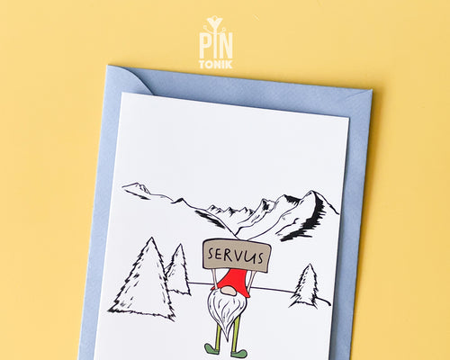 Premium postcard with an original illustration of a mountain gnome holding a 'SERVUS' sign in a snowy alpine landscape, shown with a light blue envelope.