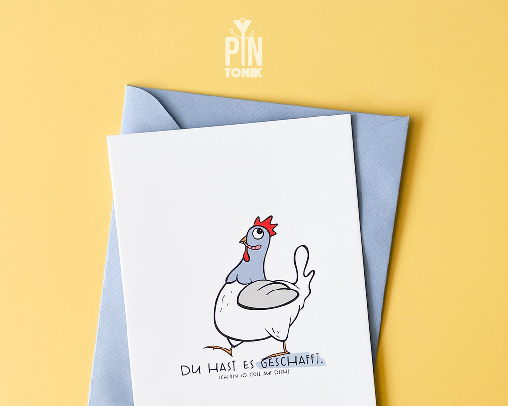 Premium postcard featuring an original hand-drawn chicken and the text 'Du hast es geschafft,' paired with a luxury blue envelope on a vibrant yellow background.