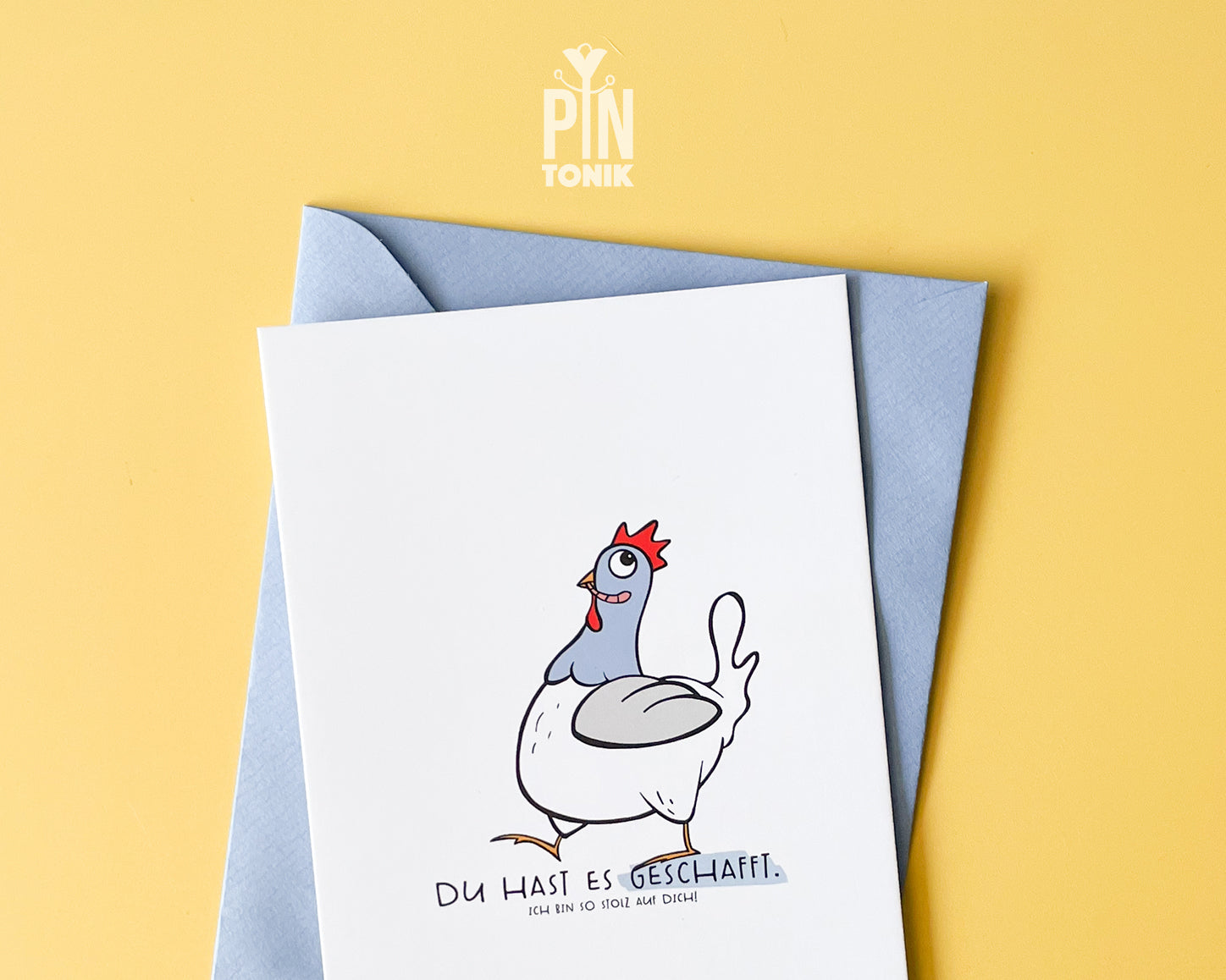 Premium postcard featuring an original hand-drawn chicken and the text 'Du hast es geschafft,' paired with a luxury blue envelope on a vibrant yellow background.