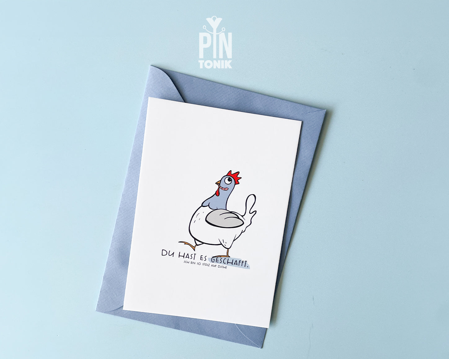 Premium postcard featuring an original hand-drawn chicken and the text 'Du hast es geschafft,' paired with a luxury blue envelope on a vibrant yellow background.