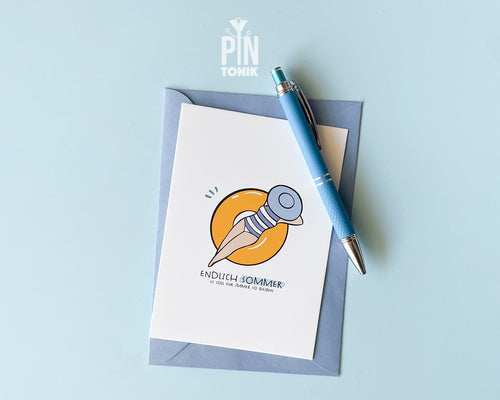 Premium postcard with an original illustration of a woman in a striped swimsuit on a yellow pool float with the text 'ENDLICH SOMMER', shown with a light blue envelope and a matching blue pen.