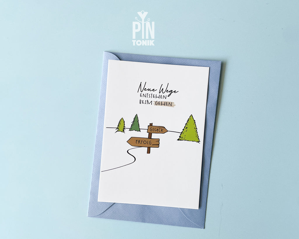 Premium postcard with an original illustration of a forest path and a signpost pointing to 'GLÜCK' and 'ERFOLG' with the text 'Neue Wege entstehen BEIM GEHEN', shown with a light blue luxury envelope.