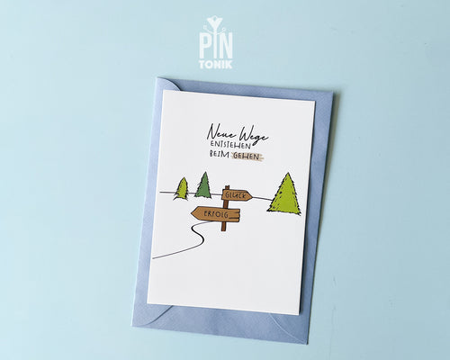 Premium postcard with an original illustration of a forest path and a signpost pointing to 'GLÜCK' and 'ERFOLG' with the text 'Neue Wege entstehen BEIM GEHEN', shown with a light blue luxury envelope.