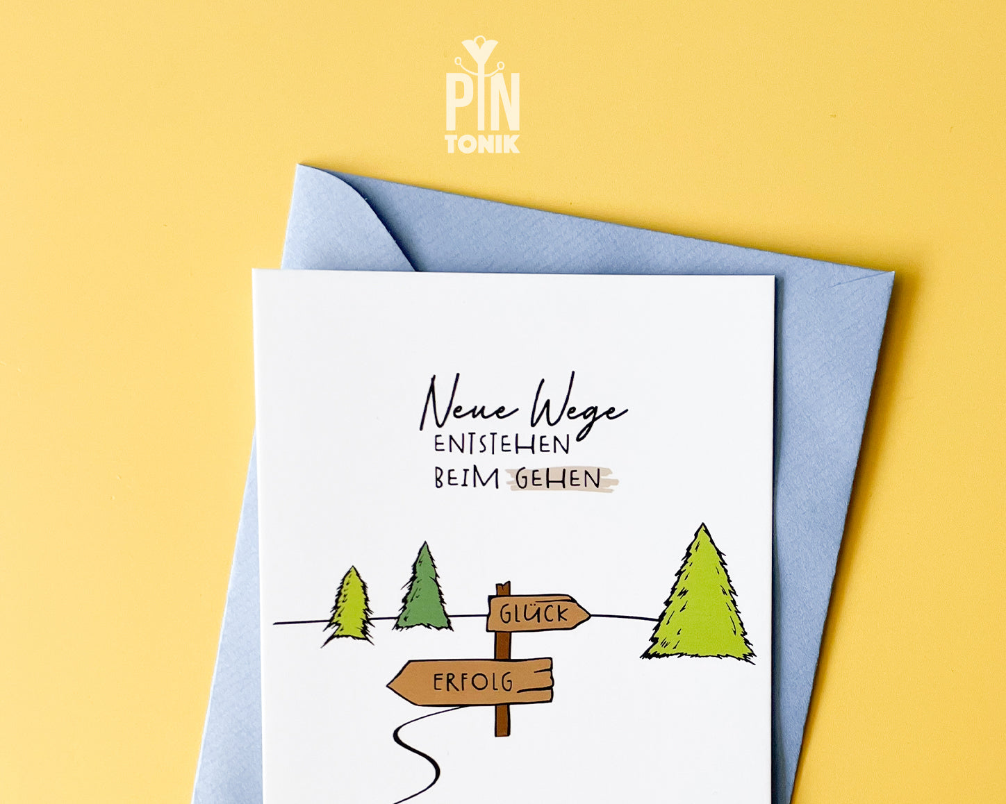 Premium postcard with an original illustration of a forest path and a signpost pointing to 'GLÜCK' and 'ERFOLG' with the text 'Neue Wege entstehen BEIM GEHEN', shown with a light blue luxury envelope.