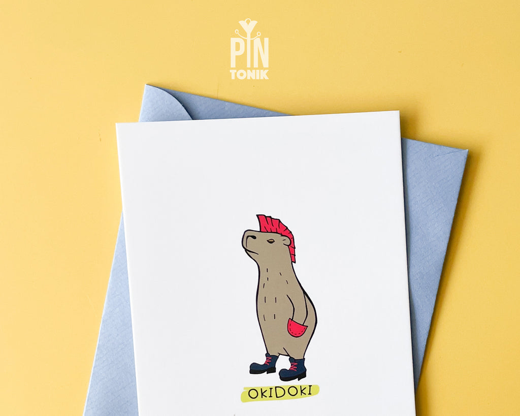 Premium postcard with an original illustration of a capybara with a pink mohawk and boots with the text 'OKIDOKI', shown with a light blue luxury envelope on yellow background.