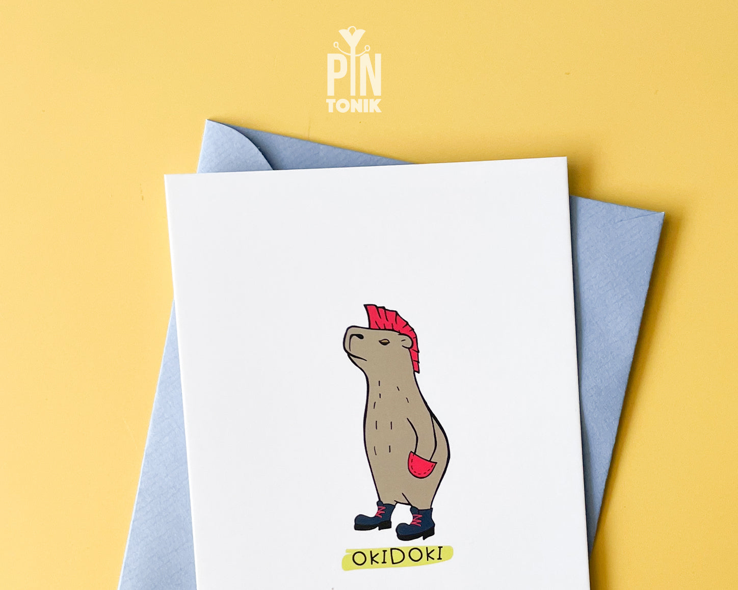 Premium postcard with an original illustration of a capybara with a pink mohawk and boots with the text 'OKIDOKI', shown with a light blue luxury envelope on yellow background.