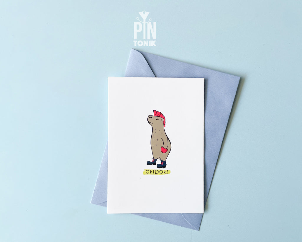 Premium postcard with an original illustration of a capybara with a pink mohawk and boots with the text 'OKIDOKI', shown with a light blue luxury envelope on a light blue background.