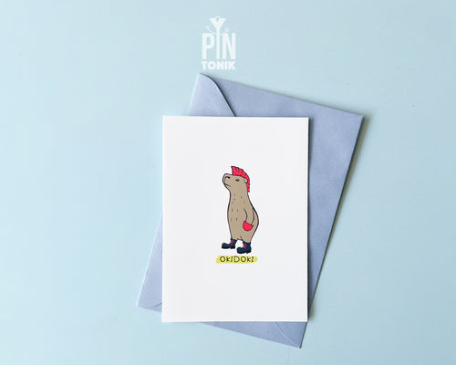 Premium postcard with an original illustration of a capybara with a pink mohawk and boots with the text 'OKIDOKI', shown with a light blue luxury envelope on a light blue background.