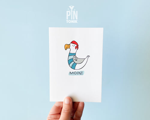 Premium postcard with an original illustration of a seagull wearing a red bandana and sailor stripes with the text 'MOIN!', shown with a light blue envelope against a soft blue background.