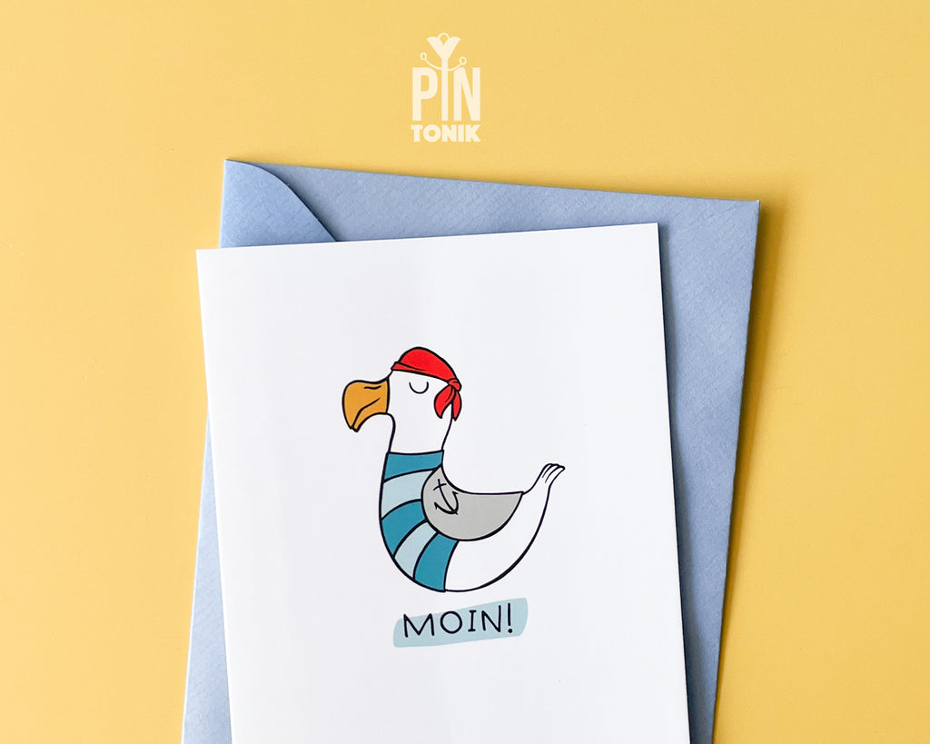 Premium postcard with an original illustration of a seagull wearing a red bandana and sailor stripes with the text 'MOIN!', shown with a light blue envelope against a soft blue background.