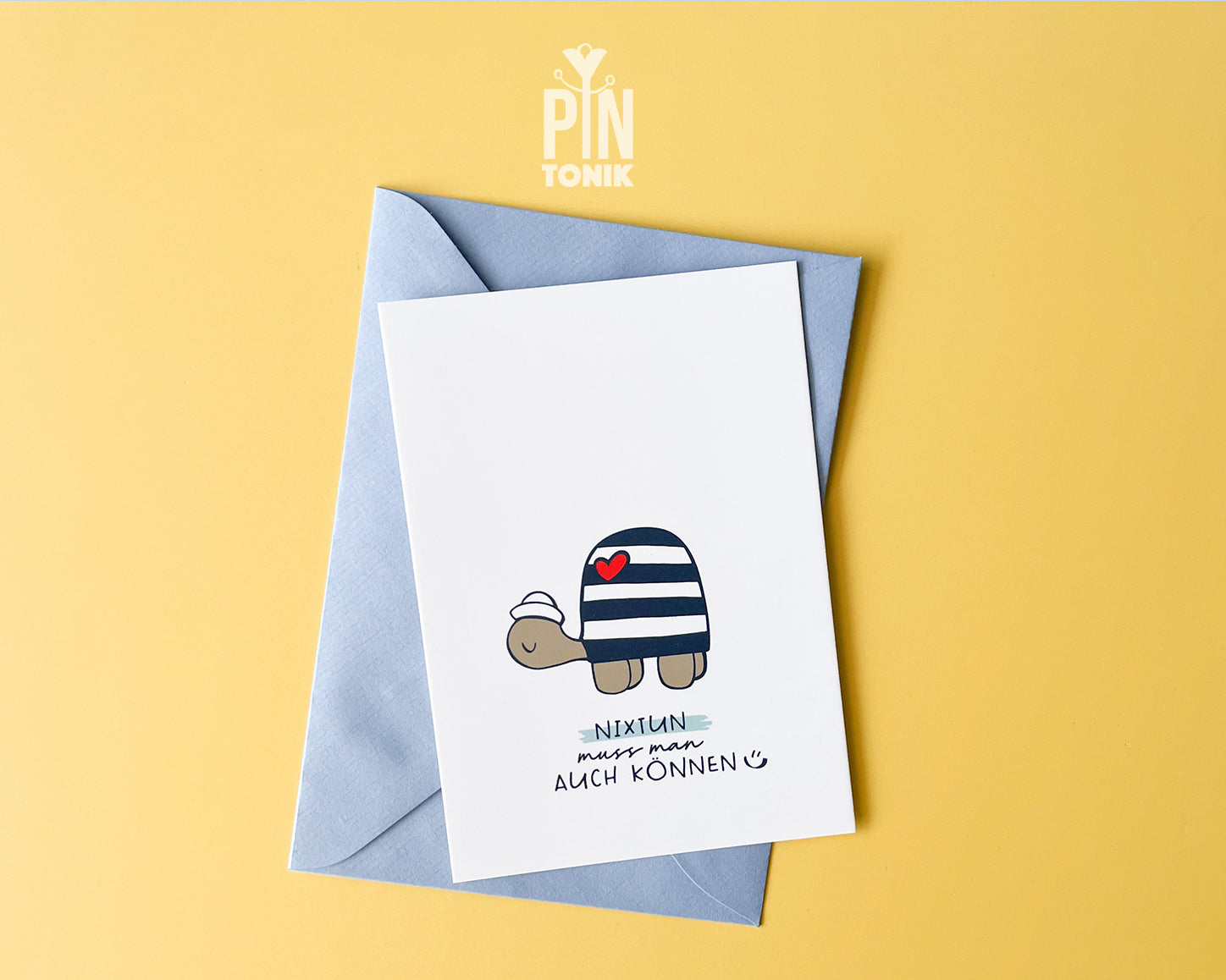Premium postcard with an original illustration of a turtle wearing a sailor hat and striped shirt with a red heart, featuring the text 'NIXTUN muss man AUCH KÖNNEN', shown with a light blue envelope.