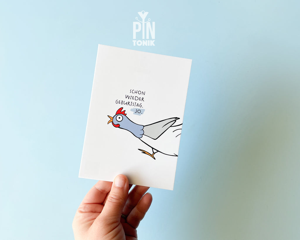 Hand holding a card with a cartoon rooster and German text on a light blue background, with 'PIN' brand visible.