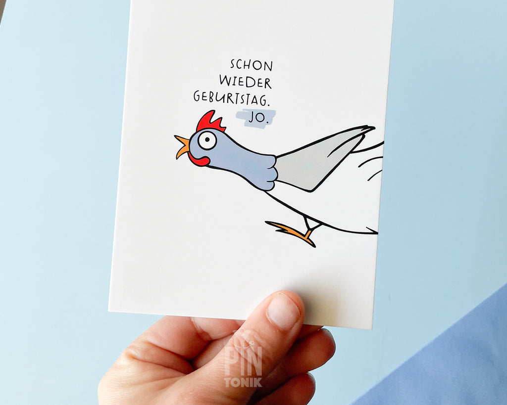 Hand holding a card with a cartoon rooster and German text on a light blue background