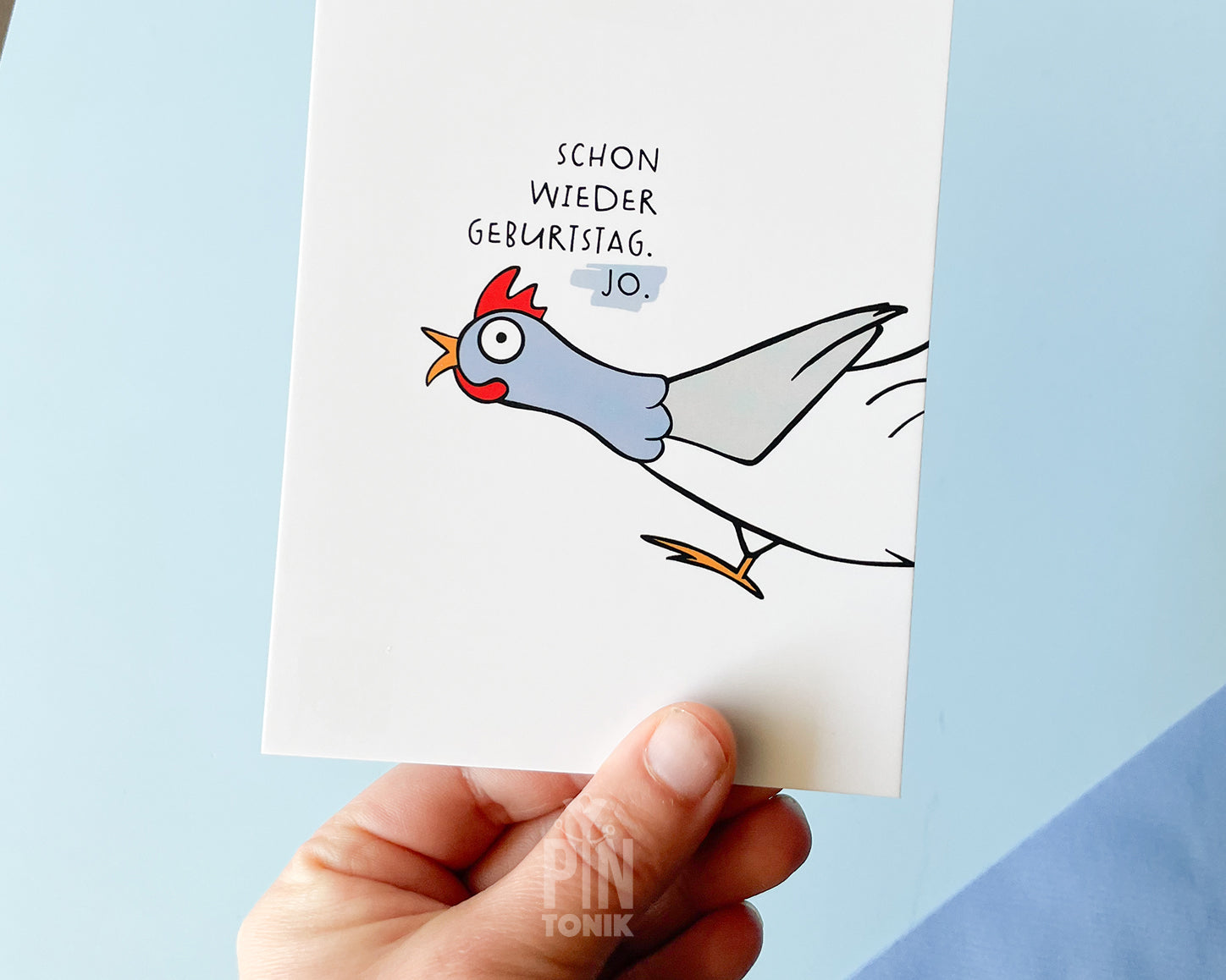 Hand holding a card with a cartoon rooster and German text on a light blue background