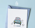 Premium postcard with an original illustration of a light blue retro car and the text 'Bon Voyage!', shown held against a light blue background.