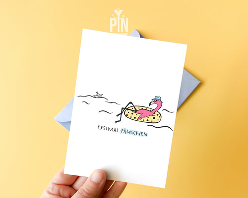 Premium postcard with an original illustration of a flamingo in a polka-dot floatie with the text 'ERSTMAL PÄUSCHEN', shown with a light blue luxury envelope.